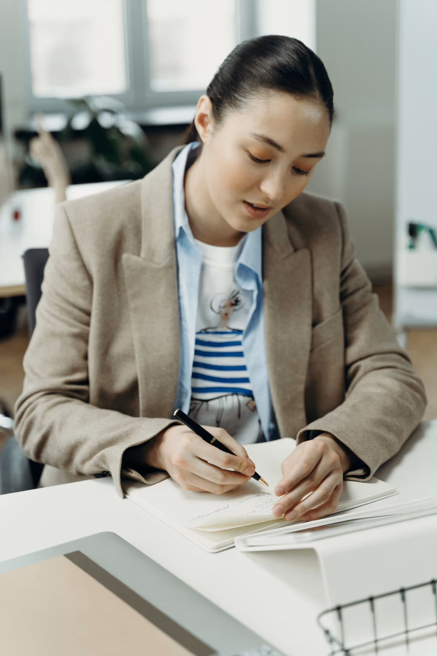 Junge Frau in einem beigen Blazer schreibt mit einem schwarzen Stift in ein Notizbuch auf einem Schreibtisch.